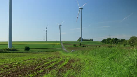 Wind-turbines-towering-over-a-lush-green-field-with-a-clear-blue-sky,-road-winding-through-the-landscape