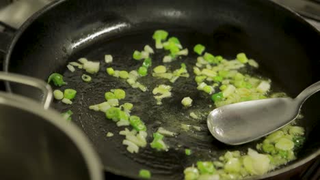 Close-up-of-chopped-onions-and-green-peppers-being-sautéed-in-a-hot-pan-with-oil,-stirred-by-a-metal-spatula,-kitchen-setting