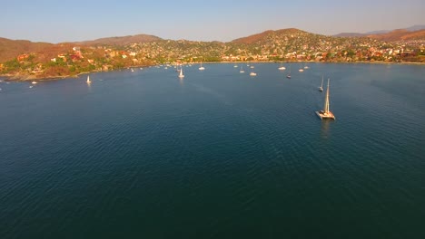 Tilting-up-to-reveal-a-busy-ocean-bay-in-Zihuatanejo,-Mexico-at-sunset-as-the-sailboat-and-yachts-return-to-anchor-near-shore