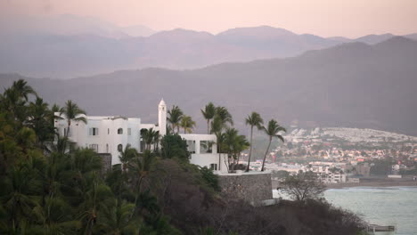 Tropical-oceanfront-villa-by-the-sea-surrounded-by-palm-trees-and-beautiful-mountains-in-the-distance-at-dawn
