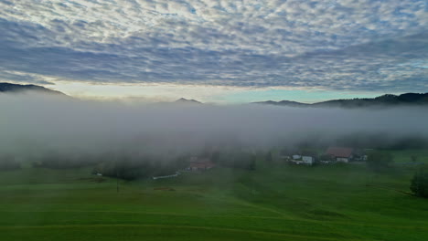 Nebliges-Tal-Bei-Sonnenaufgang-Mit-Wolken-über-Dem-Kopf-Und-Verstreuten-Häusern,-Luftaufnahme