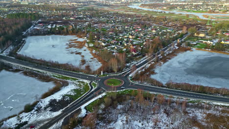 Frozen-lake-next-to-a-road-with-roundabout,-early-winter,-houses-in-the-background,-aerial-view
