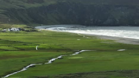 Kiteboarding-Down-Narrow-River-Near-Rough-Ocean-Waves,-Green-Meadows,-Lush-Green-Beautiful-Landscape,-Achill-Island,-Ireland