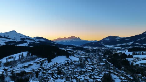 Die-Drohne-Fliegt-über-Ein-Dorf-In-Einem-Schneebedeckten-Tal-Mit-Großen-Bergen-Im-Hintergrund-Und-Dem-Sonnenuntergang-In-Österreich,-Luftaufnahmen-4K