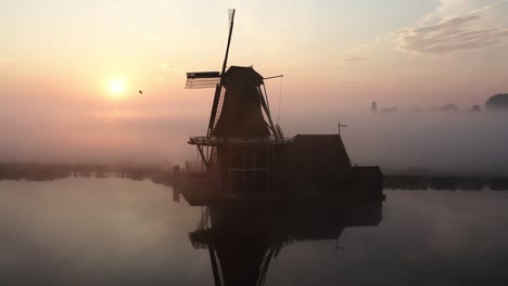 Die-Drohne-Fliegt-Um-Eine-Windmühle-Herum,-Während-Die-Aufgehende-Sonne-Hinter-Der-Windmühle-Sichtbar-Wird-Und-Viel-Nebel-In-De-Zaanse-Schans,-Den-Niederlanden,-Luftaufnahmen-In-4K