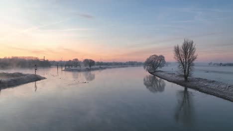 Panoramic-drone-shot-with-a-cargo-ship-passing-by-on-the-ijssel-on-an-ice-cold-foggy-morning-with-a-beautiful-sunrise