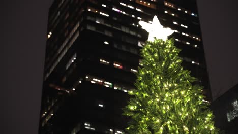 Close-up-of-the-tip-of-a-very-large-green-simple-christmas-tree-with-a-golden-glowing-star-on-top-with-the-lights-of-city-skyscrapers-in-the-background-at-night-in-winter