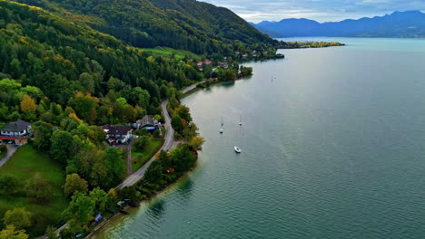 Serene-lakeside-with-sailboats,-bordered-by-lush-greenery-and-houses,-under-a-cloudy-sky,-aerial-view