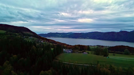Serene-lake-surrounded-by-hills-and-a-forested-landscape-at-dusk,-clouds-overhead,-aerial-view