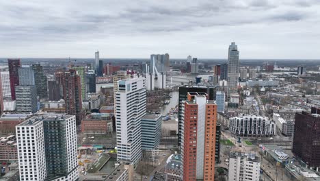 Die-Drohne-Fliegt-über-Hohe-Gebäude-Mit-Der-Erasmus-Brücke-Im-Hintergrund-An-Einem-Bewölkten-Tag-In-Rotterdam,-Niederlande.-Luftaufnahmen-In-4K