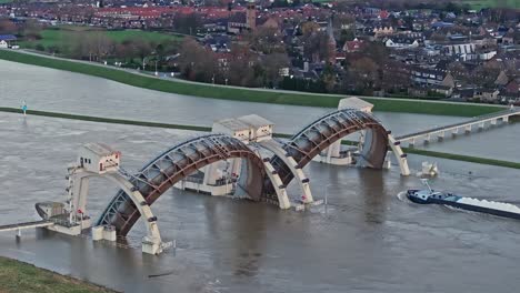 Zoomed-in-drone-shot-moving-towards-the-weir-of-Driel-with-the-town-of-Driel-in-the-background-during-high-water-levels-with-the-doors-open-and-a-cargo-ship-passing-thru