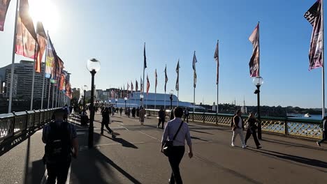 People-walking-across-Pyrmont-bridge-Darling-harbour-Sydney,-with-bright-sun-and-flags-blowing-in-the-breeze