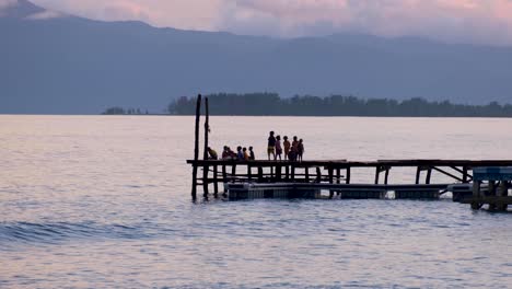 Children-watching-beautiful-sunset-on-wharf-overlooking-ocean-water-on-a-remote-tropical-island-in-Raja-Ampat,-West-Papua-in-Indonesia