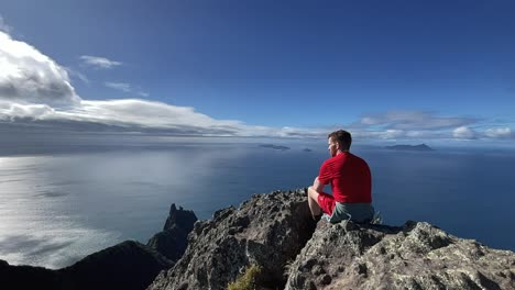 Male-sits-on-mountain-top,-looks-out-at-ocean-and-blue-sky-clouds,-New-Zealand