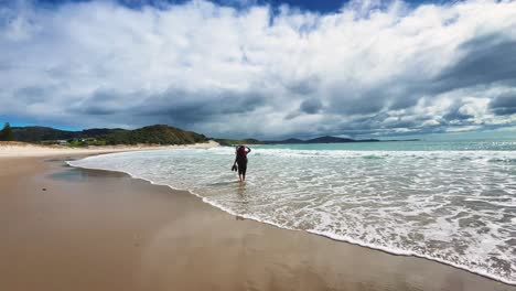 Backpacker-wades-through-surf-at-beach-on-cloudy-day,-slow-motion-wide-shot