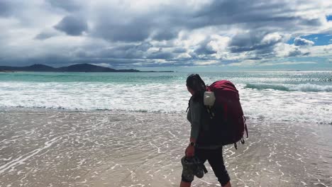 Asian-female-backpacker-walks-in-ocean-waves-at-bright-cloudy-beach