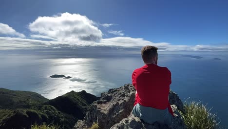 Rothaariger-Männlicher-Wanderer-Blickt-Auf-Das-Meer-Mit-Flauschigen-Weißen-Wolken,-Neuseeland