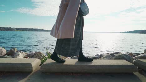 woman-in-coat-walking-along-the-lakeside-at-the-sunset-in-fall-evening-slow-side-walk-middle-shot-close-up-leg