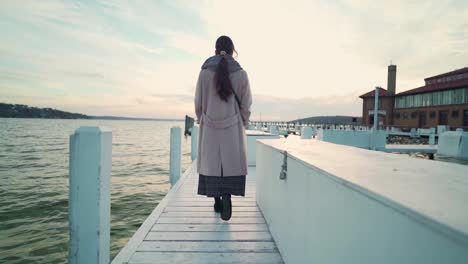 Asian-woman-walking-along-the-dock-towards-the-lake-water-at-sunset-in-chilly-autumn-evening-slow-motion