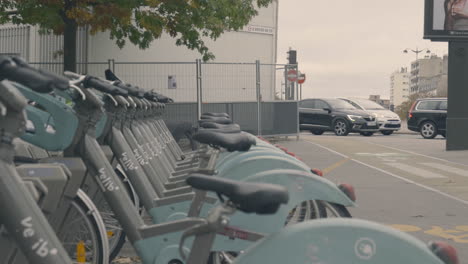 Velib-Electric-City-Bicycles-Parked-in-Line,-Cars-passing-in-the-Background