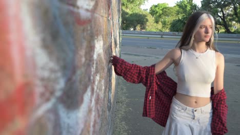 Beautiful-young-woman-walking-worried-dragging-her-hand-on-wall-of-park-at-sunset