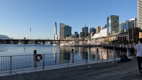 Couple-walking-along-the-boardwalk-at-Darling-Harbour-Sydney,-pan-left-with-tall-city-buildings