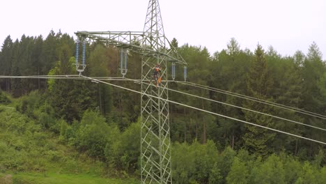 Worker-climbing-a-high-voltage-power-tower-amidst-green-forest,-overcast-day,-aerial-shot