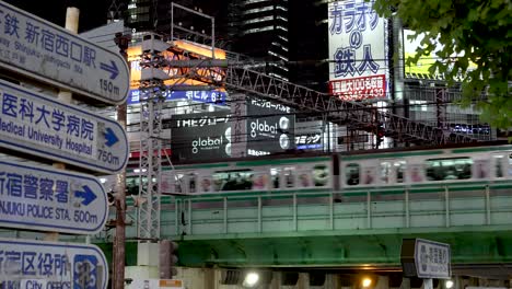 Der-Zug-Der-Yamanote-Linie-Fährt-Nachts-Auf-Der-Hochbahnstrecke-In-Shinjuku-Vorbei-Und-Zeigt-Informationsverkehrsschilder-Im-Blick