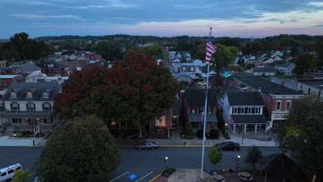 Dusk-view-over-a-town-square-with-an-American-flag,-surrounded-by-autumn-trees
