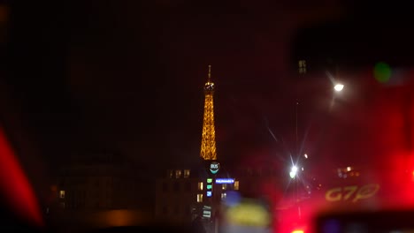 View-of-Eiffel-tower-at-night-from-inside-taxi-cab-with-motorcyclists-passing-by-while-stopped,-Handheld-view