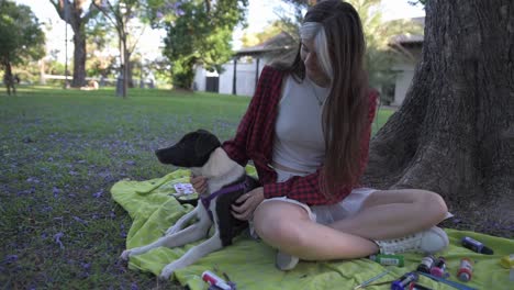 Charming-young-woman-caressing-her-dog-sitting-under-tree-surrounded-by-paintings-and-book-at-sunset