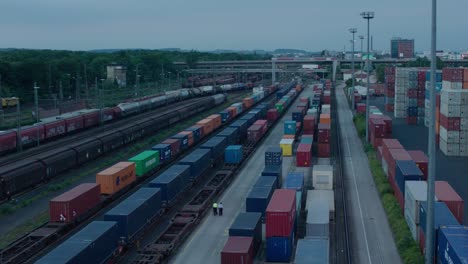 Dämmerung-über-Einem-Belebten-Güterbahnhof-Mit-Sicherheitsgängen-Und-Zahlreichen-Bunten-Frachtcontainern