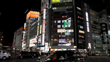 In-the-nocturnal-ambiance-of-Shinjuku,-Tokyo,-Japan,-individuals-stroll-alongside-bustling-car-traffic,-contributing-to-the-vibrant-and-dynamic-atmosphere-of-the-metropolitan-night