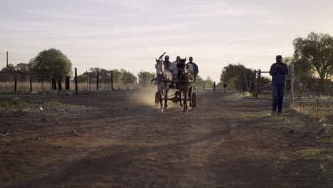 Driver-and-passengers,-carties,-ride-horse-and-scrap-metal-cart-at-sunset-down-rural-dirt-road-in-township-of-South-Africa