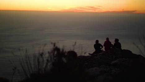 Three-young-friends-are-silhouetted-while-drinking-sundowners-on-top-of-a-mountain-during-a-beautiful-orange-sunset-with-rolling-clouds