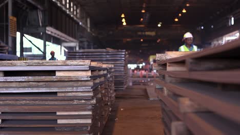 Gelagerte-Gestapelte-Stahlplatten-Im-Lagerhaus-Des-Stahlwerks,-Im-Hintergrund-Laufen-Stahlarbeiter