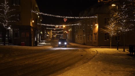 Christmas-holiday-lights-in-a-small-town-on-a-snowy,-winter-night-and-a-vehicle-driving-by