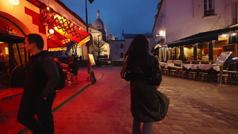 Young-woman-spinning-and-smiling-while-walking-down-a-street-with-restaurants,-in-Paris-near-Montmartre,-at-night