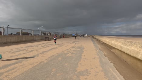 Charity-runners-running-along-a-seafront-next-to-a-sandy-beach-with-grey-storm-clouds-and-bright-sunshine