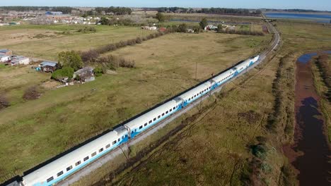 Train-gliding-along-tracks-near-a-winding-river-with-rural-homes-in-the-backdrop,-on-the-outskirts-of-Buenos-Aires