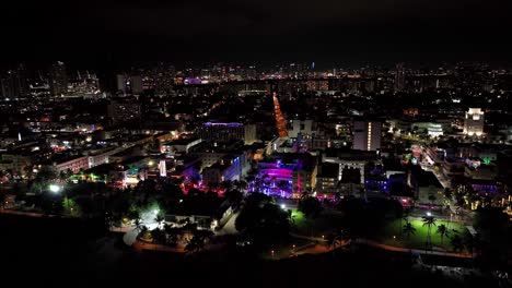 Suggestive-nightly-aerial-drone-footage-of-Miami,-USA:-slow-backwards-movement,-from-the-city-with-buildings,-colored-lights,-roads,-to-the-seaside-and-the-darkness-of-the-sea