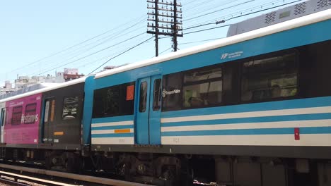 Slow-Motion-Public-Blue-and-White-Train-Drives-Through-Rails-in-Summer-Skyline-at-Sarmiento-Line,-Flores-Station