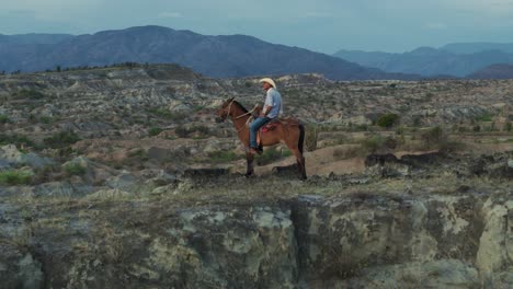 Old-Fashioned-Cowboy-on-Horseback-in-Majestic-Tatacoa-Desert-Landscape,-Aerial-Reveal
