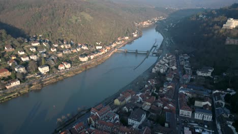 Aerial-view-of-a-picturesque-European-town,-alongside-a-river,-at-the-base-of-a-mountain,-on-a-sunny-day,-featuring-a-bridge-and-views-of-the-village-and-lush-mountains