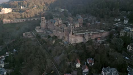 Aerial-view-of-a-historic-castle-on-a-hillside,-overlooking-a-village,-surrounded-by-a-forest-of-trees