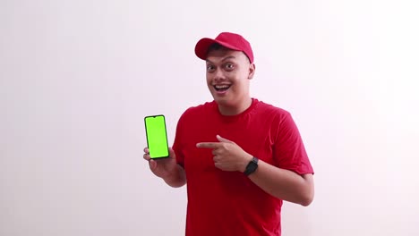 Young-handsome-asian-courier-man-showing-green-screen-phone-display-and-pointing-on-it,-making-thumbs-up-hand-gesture