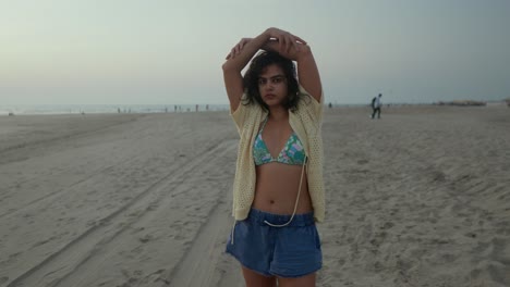 Young-woman-in-a-bikini-top-and-open-cardigan-at-the-beach-during-dusk,-arms-raised,-casual-and-relaxed
