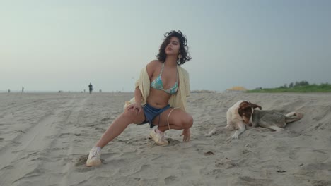 Young-woman-in-a-playful-pose-on-a-sandy-beach-with-a-dog-lying-next-to-her,-casual-beachwear,-overcast-day