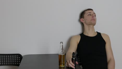 Young-depressed-man-sitting-alone-at-the-table-with-a-beer-and-smoking,-vaping-inside