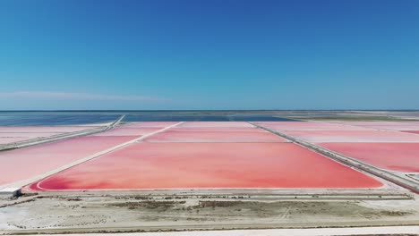 Flying-above-Albanian-sea-salt-farm-with-pink-sediment,-blue-sky-seaside